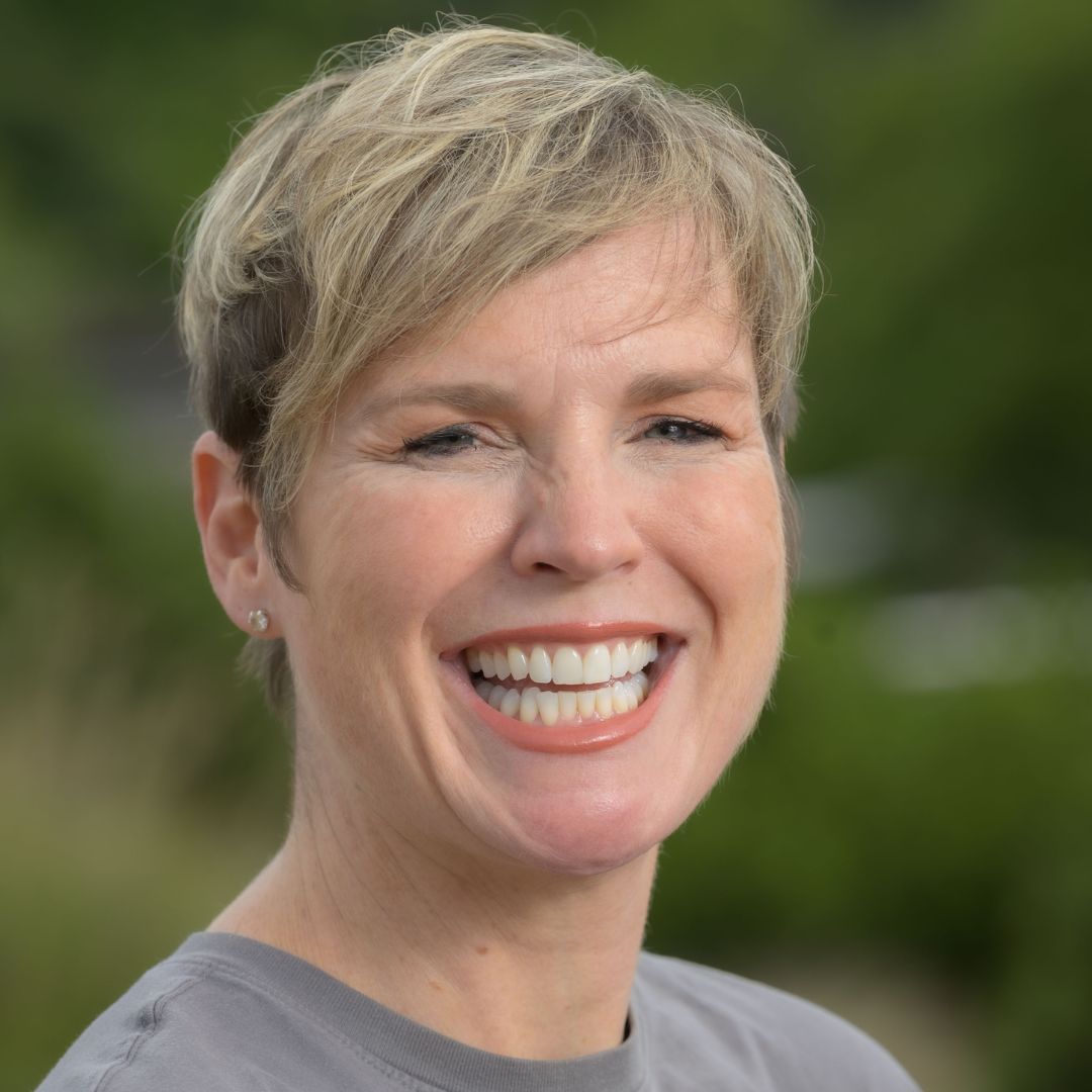 Headshot of Dani Carlaw who wears a grey top and stands in front of a green background.