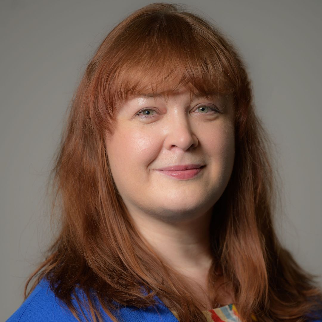 Headshot of Louise Acheson, who wears a royal blue top and stands in front of a grey background.