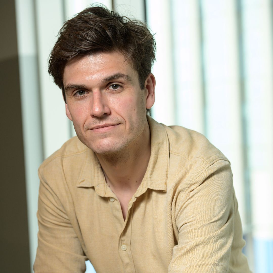 A headshot of Sean, who leans forward on a desk and wears a beige shirt.