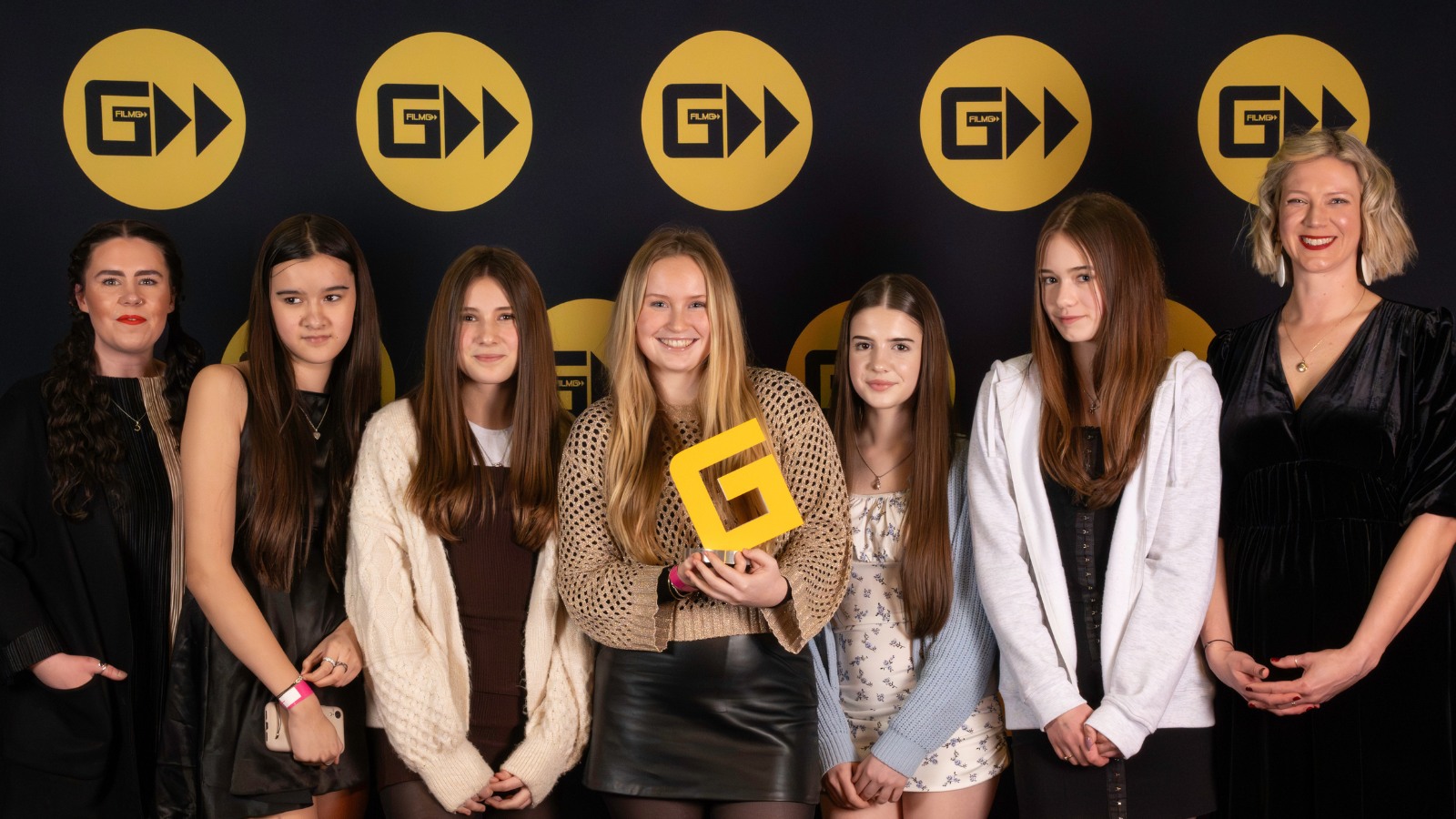 A group of girls and women stand in a line, with the person in the middle holding a trophy in the shape of the letter G