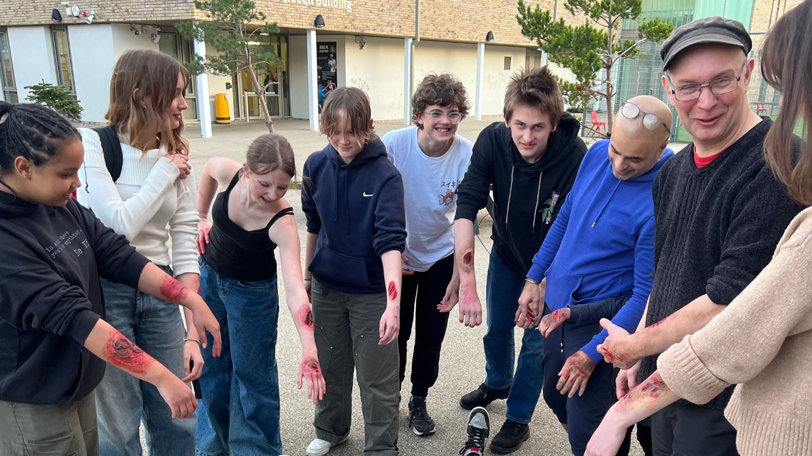 A group of school pupils stand in a line outside in a school playground. They hold out an arm each, which all have special effects make up on them, in a range of wounds and cuts.