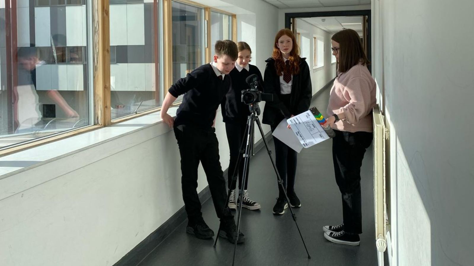 Photo taken at Lochaber High School of a FilmG club - four pupils stand around a camera on a tripod in a school corridor. One holds a clapper board which reads FilmG on it.