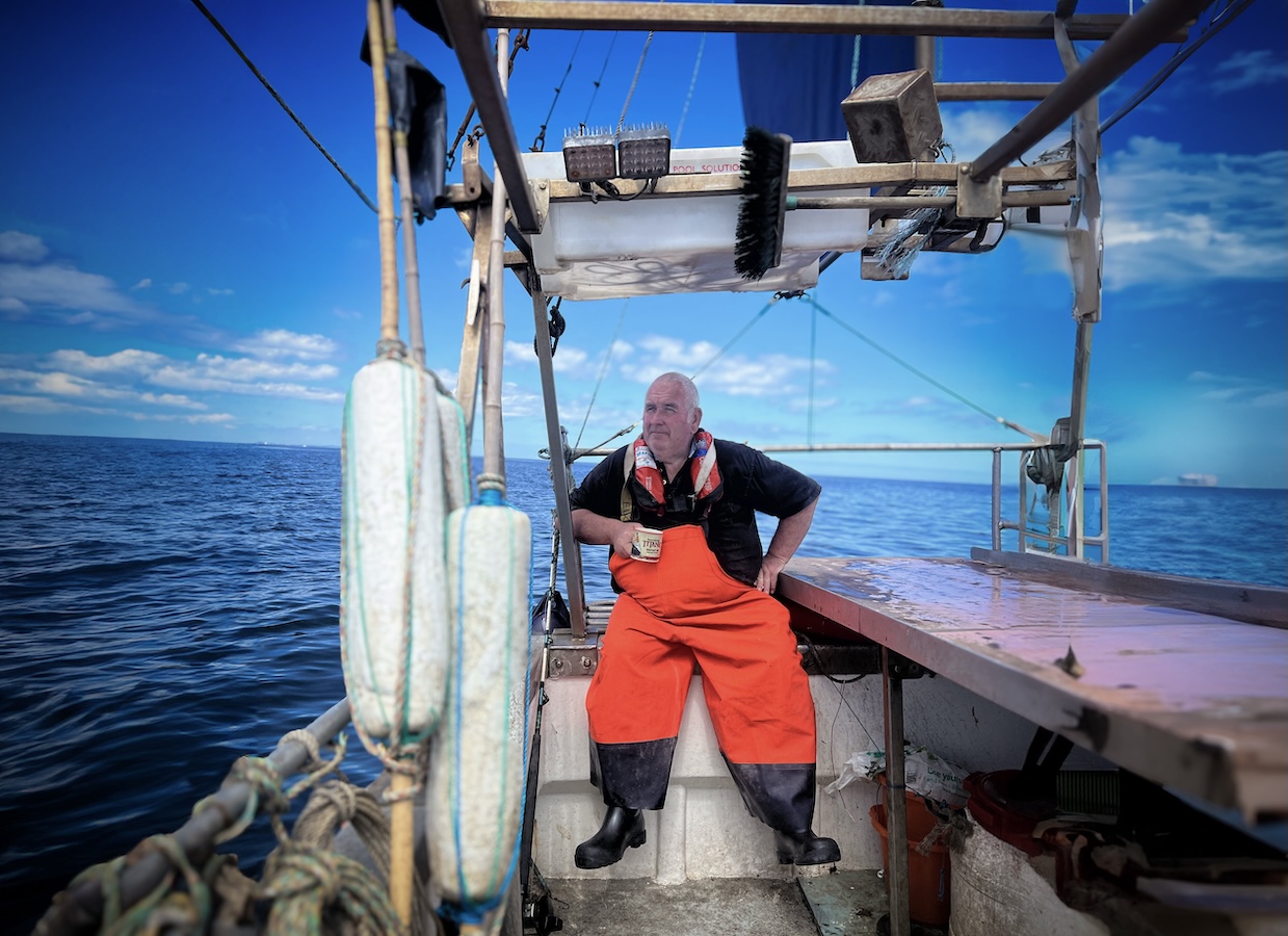 A man wearing bright orange overalls sits on a wooden boat, holding a mug. The sky is clear and blue, with some fluffy clouds, and the calm sea stretches out in the background. Equipment and ropes are visible on the boat.