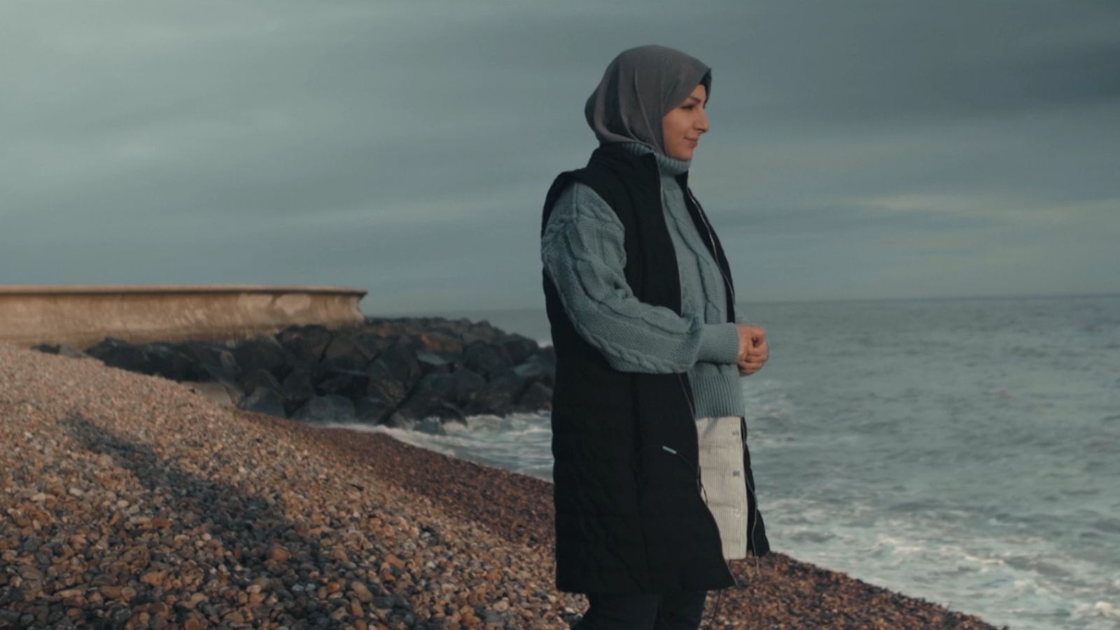 A person wearing a hijab and a dark vest over a light grey sweater stands on a pebbled beach, gazing out at the sea under an overcast sky.
