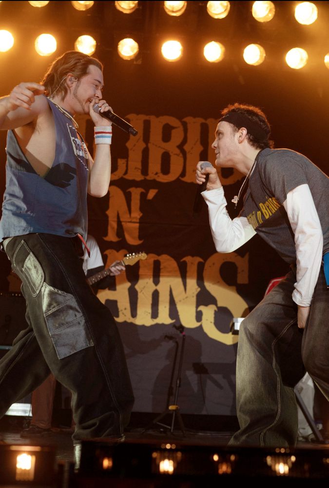 Still from California Schemin', which shows actors Samuel Bottomley and Séamus McLean Ross on a stage wearing 90s clothing. They each hold a microphone and are rapping into these.