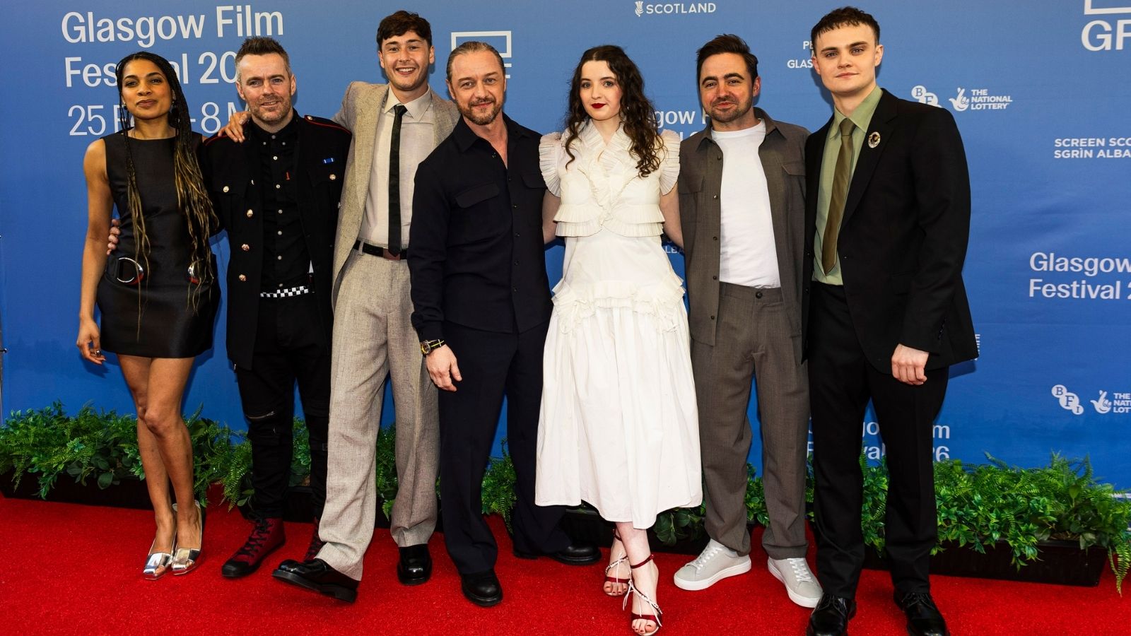 A group of seven people stand on a red carpet in front of a blue backdrop at the Glasgow Film Festival. They are dressed in a variety of outfits, from sleek dresses to smart suits. Green foliage lines the edge of the carpet. The backdrop features festival and sponsor logos.