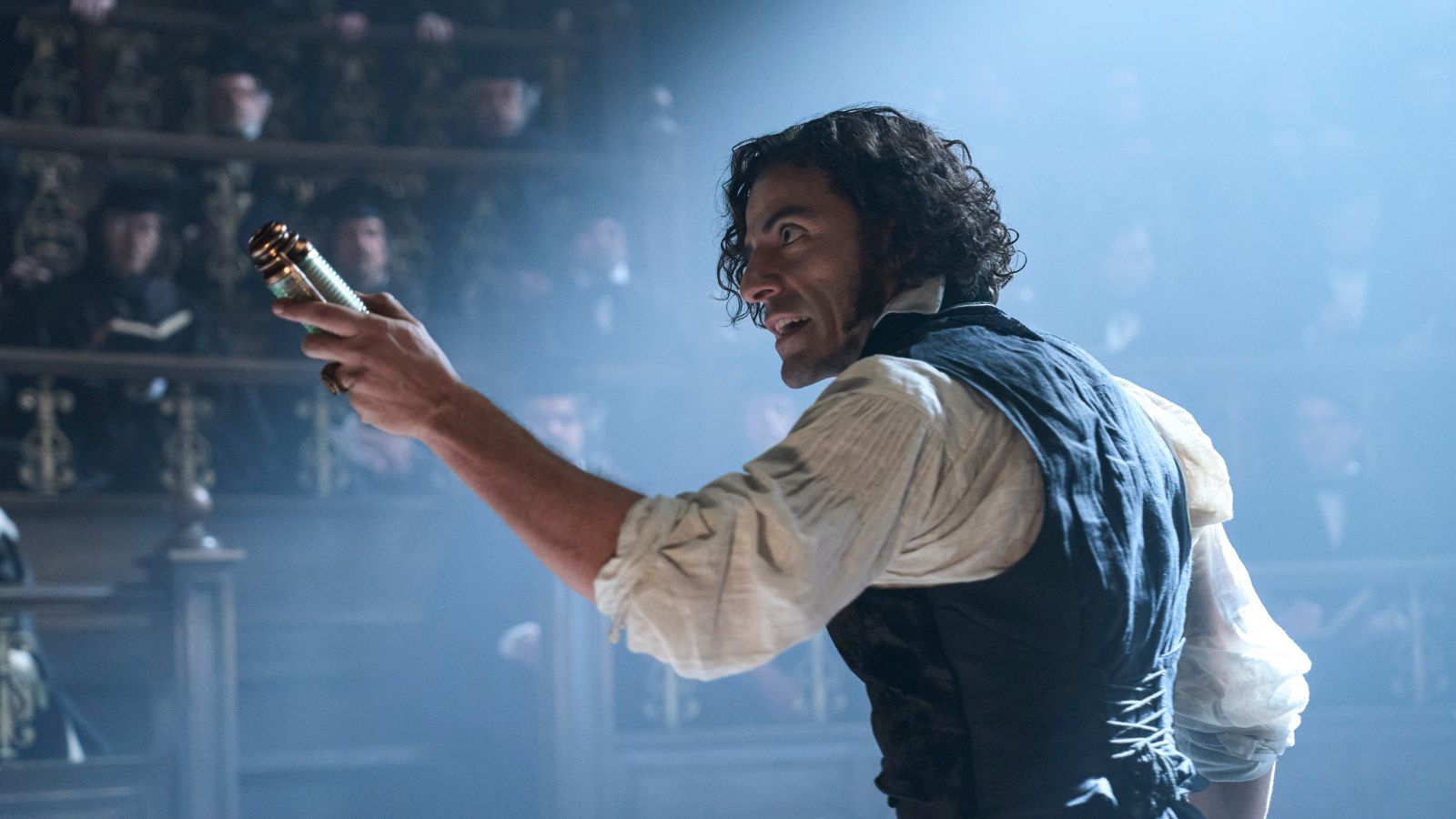 Still from Frankenstein, which shows actor Oscar Isaac as Dr Victor Frankenstein, wearing a white shirt and a waistcoat. He holds up a scroll and is addressing rows of men who watch him speak.