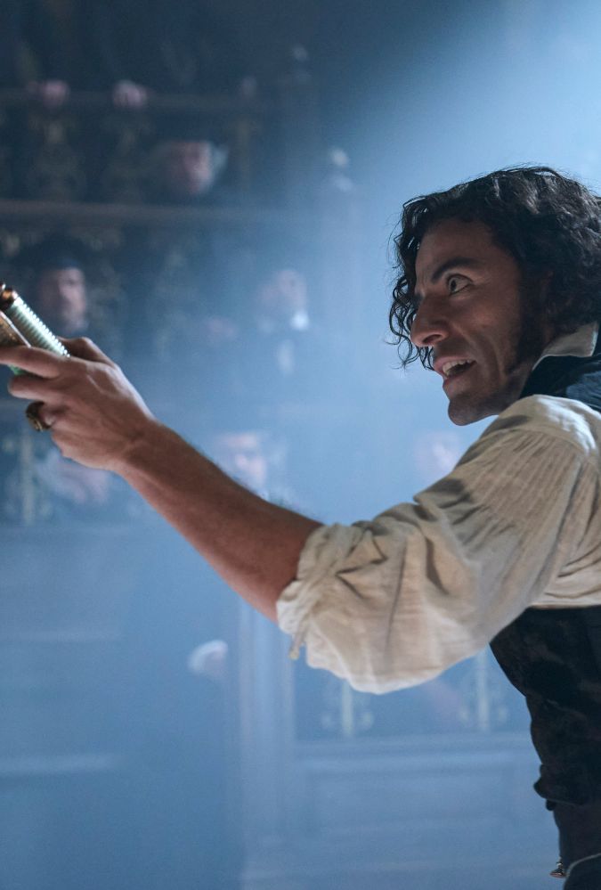 Still from Frankenstein, which shows actor Oscar Isaac as Dr Victor Frankenstein, wearing a white shirt and a waistcoat. He holds up a scroll and is addressing rows of men who watch him speak.