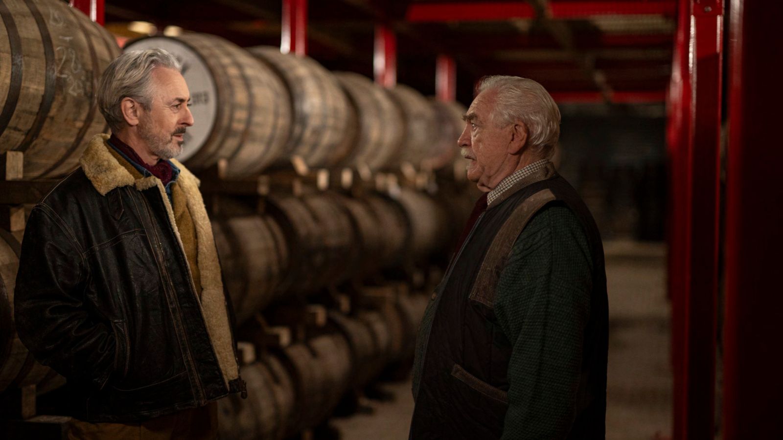 Still from Glenrothan, which shows actors Alan Cumming and Brian Cox standing facing each other inside a distillery with wooden whiskey barrels stacked up in rows behind them