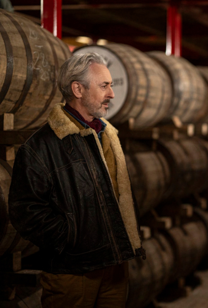 Still from Glenrothan, which shows actor Alan Cumming inside a distillery with wooden whiskey barrels stacked up in rows behind him.