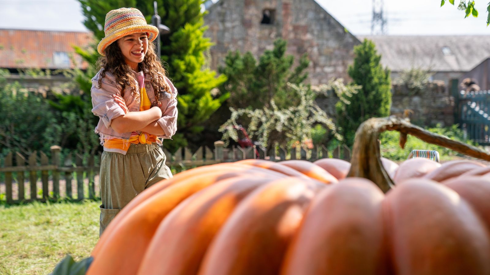 A young person with long curly hair, wearing a colourful straw hat and casual clothes, stands smiling with arms crossed, gazing at a large orange pumpkin in a garden setting. A rustic building and green foliage are in the background.