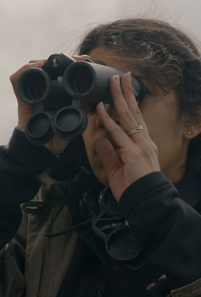 Still from Lost For Words shows a close up of a woman outdoors holding a pair of binoculars up to her face to look through them