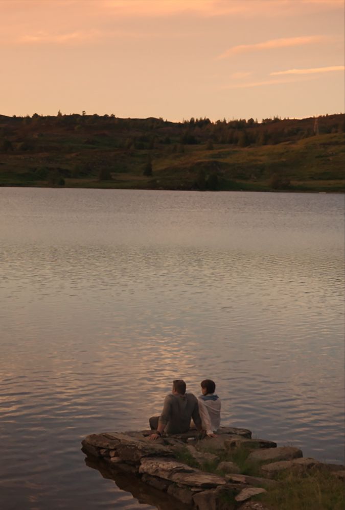 Still from Love and Trouble, showing a two people sitting outside by a loch at sunset