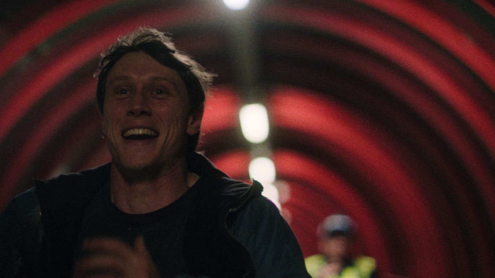 A man runs with a joyful expression in a tunnel illuminated by red and white lighting. There is another person blurred in the background.