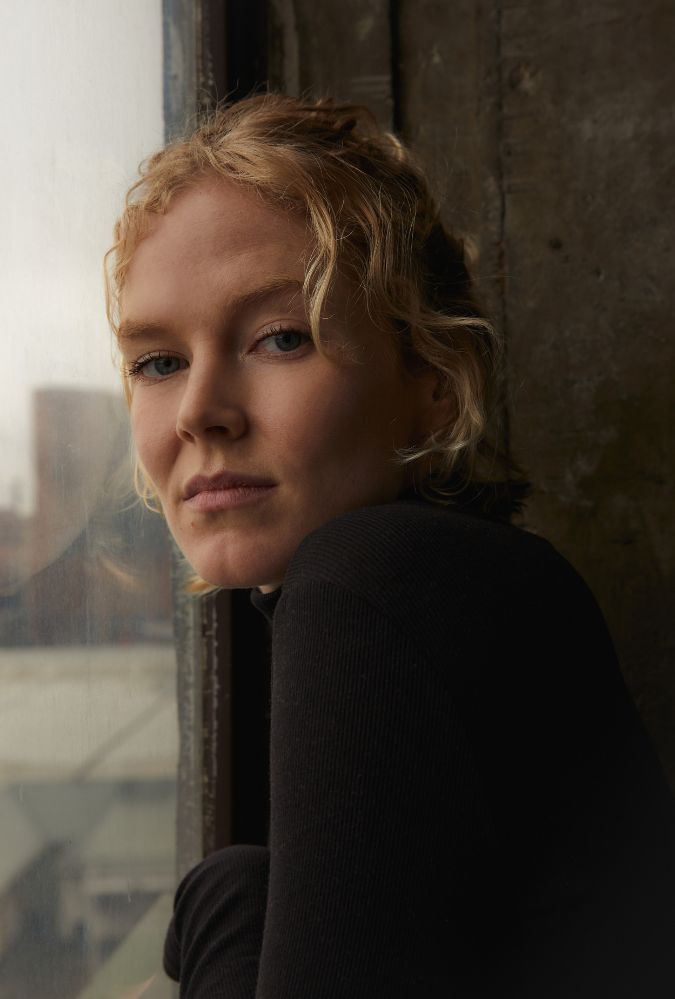 Headshot of actress Rosy McEwen, who looks at the camera, a window to her left. She wears a black top.  Photo Credit: Klara Waldberg