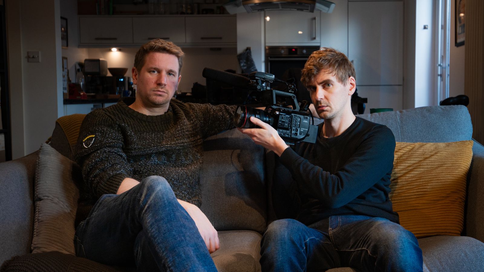 Still from Silent Men. Two men sit side by side on a sofa indoors. The man on the right (director Duncan Cowles) holds a camera and is pointing it towards the man on the left.