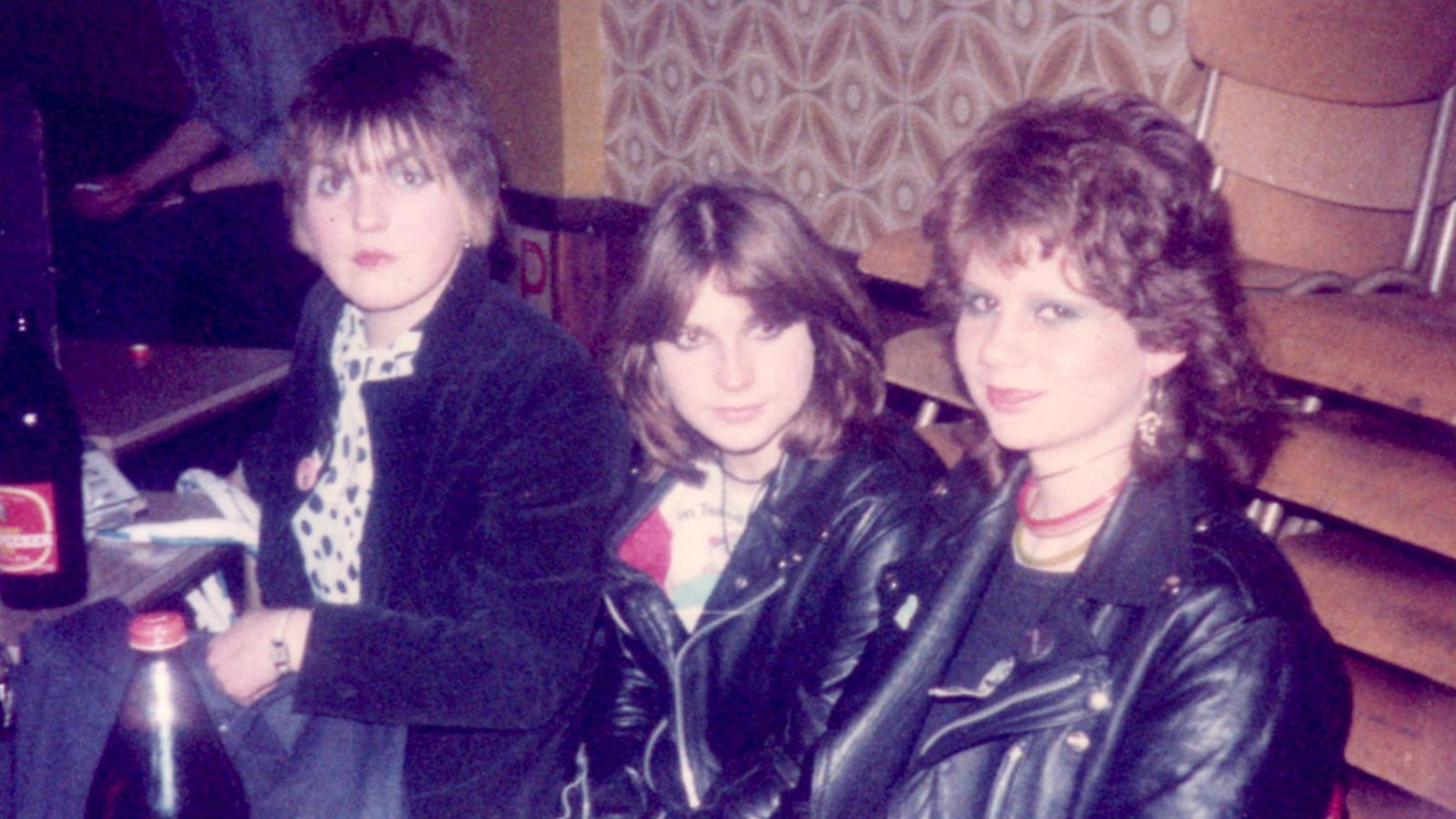 Image of band The Ettes. Three women sit in a bar area, waring leather jackets and have 1980s hair and makeup.