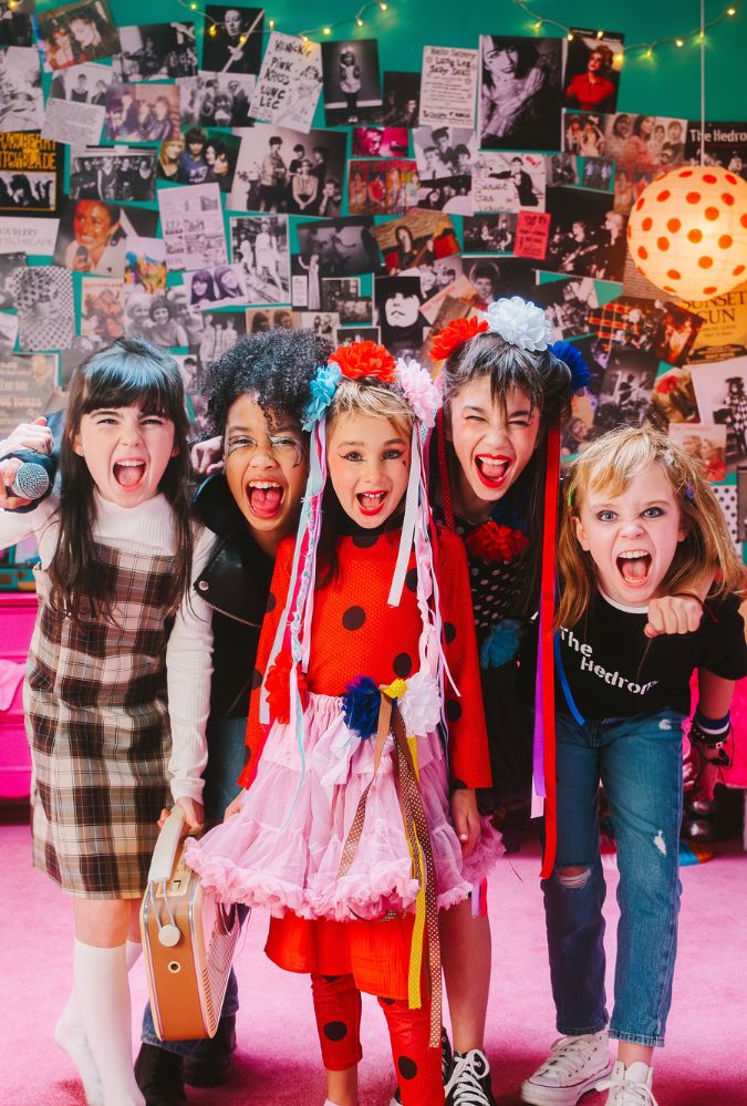 The image shows five girls in a bedroom, which is decorated in posters and images of girl groups from over the years. The girls stand in a row, yelling at the camera, wearing a range of clothing such as a plaid dress, leather jacket, jeans and t-shirt.