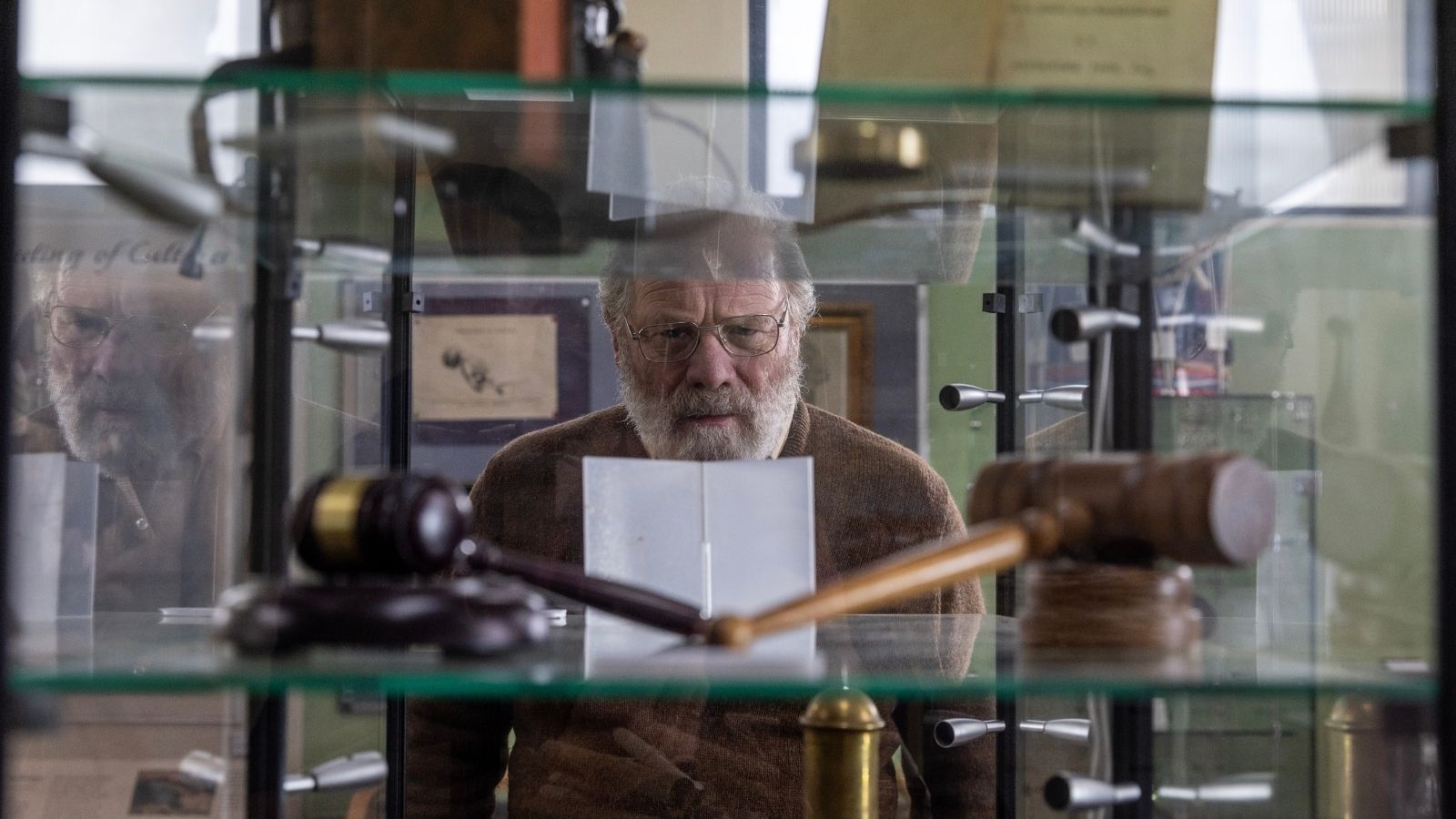 An older man with a beard and glasses intently examines a display case filled with various objects, including a wooden gavel. The setting appears to be a museum or historical exhibit.