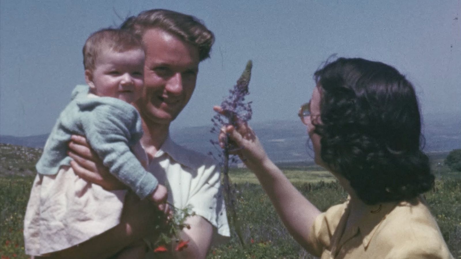 Still from The Flowers Stand Silently, Witnessing showing a man holding a child, while a woman to their right holds up a wildflower. They stand outdoors in a sunny location.