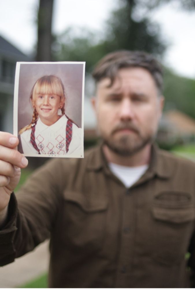 Still from The Secret of Me, which shows a man standing outdoors, holding a photograph of a young girl out in front of him.