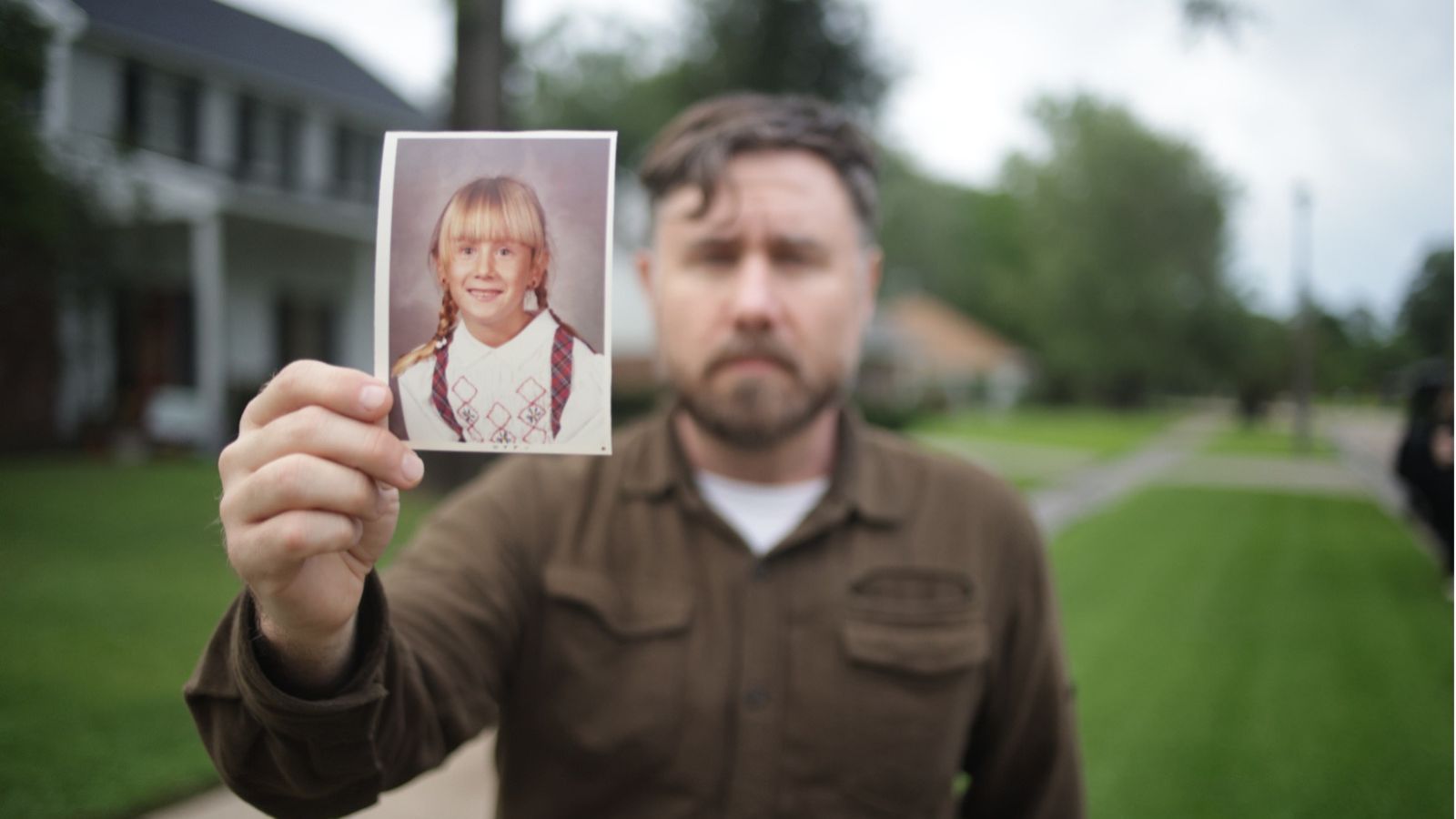 Still from The Secret of Me, which shows a man standing outdoors, holding a photograph of a young girl out in front of him.