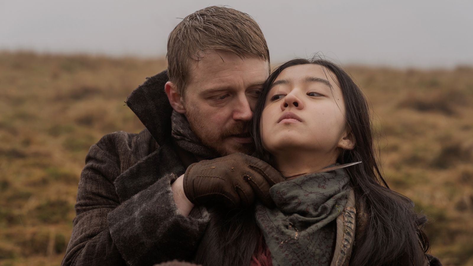 Still from Tornado, which shows actor Jack Lowden holding a knife to actor Koki's neck. They are outdoors in a rural setting wearing clothing from the 1790s.