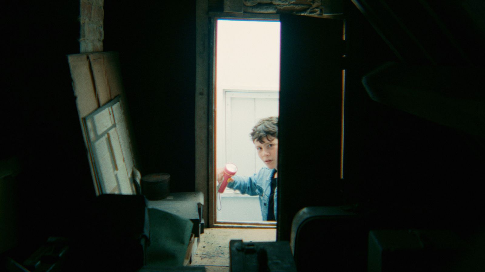 Still from Where Dragons Live, courtesy of Bombito Productions, showing a boy looking through a door into a shed. He holds. torch and wears a blue denim jacket.