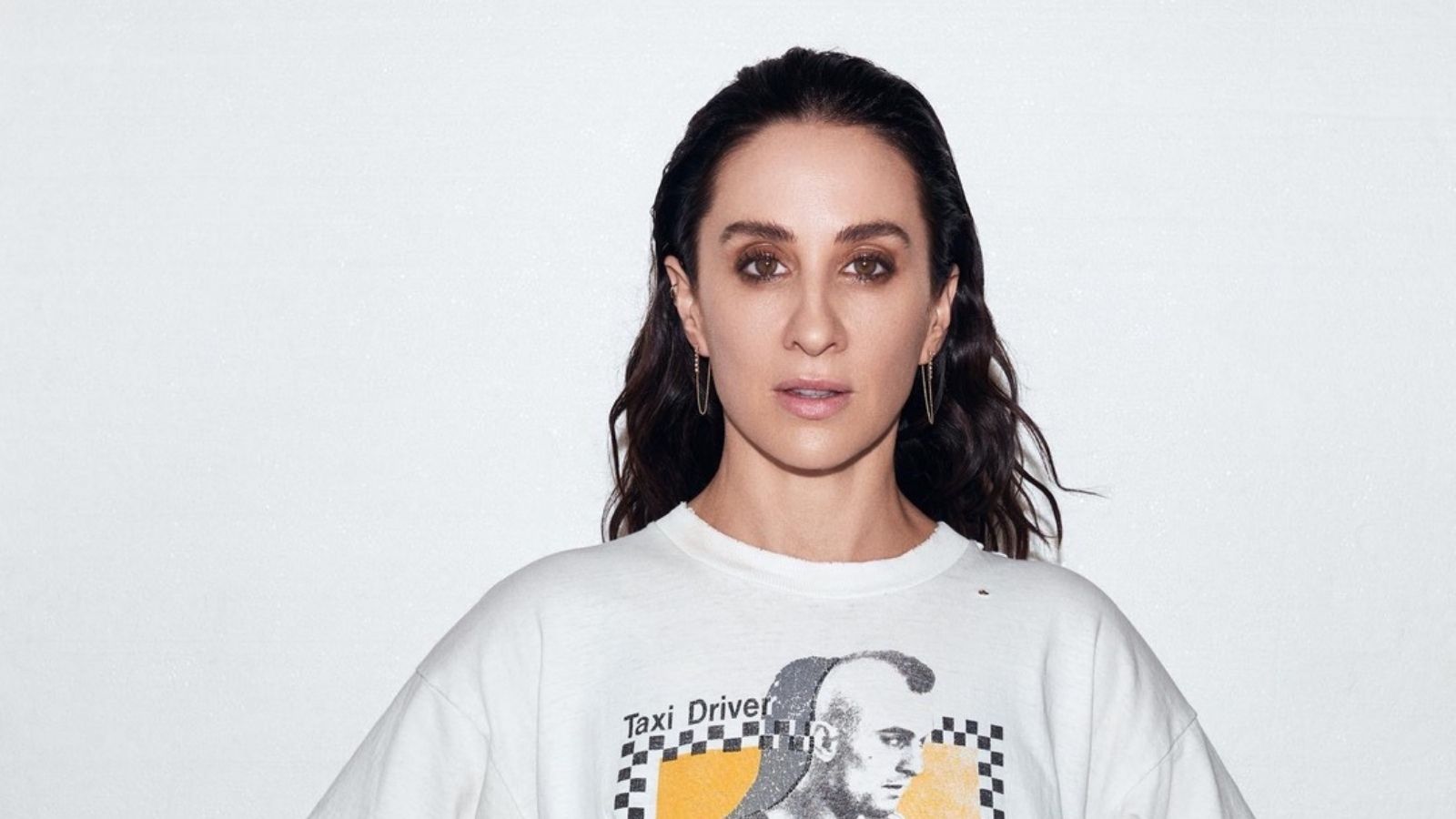 A person with dark hair wearing a white T-shirt featuring a graphic of a man with a mohawk and the phrase "Taxi Driver." The background is plain white.