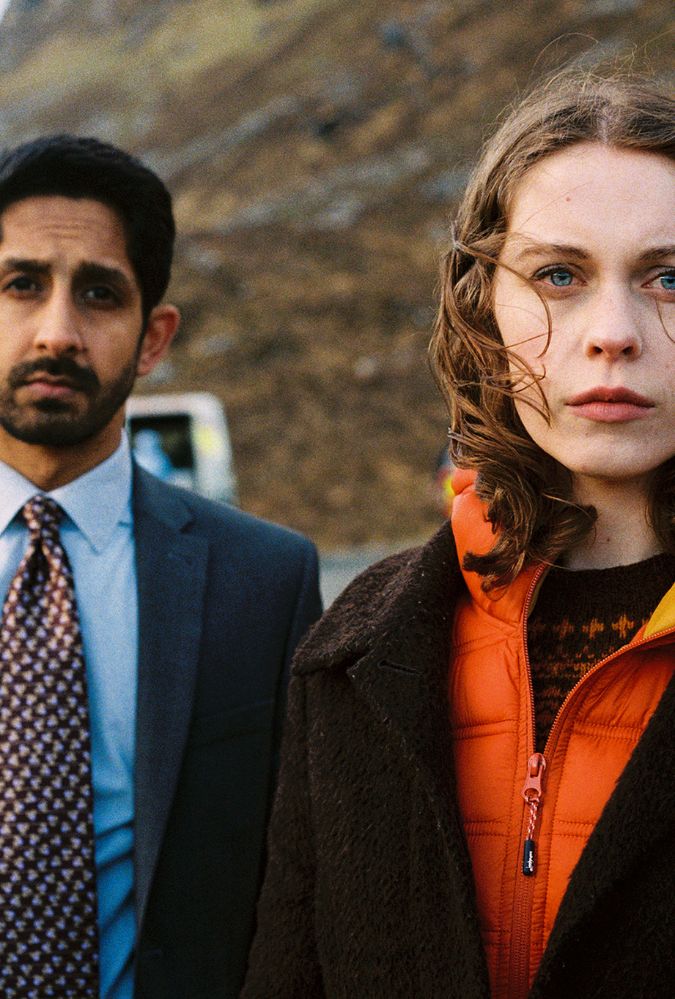 Still from An t-Eilean (The Island), which shows actor Sagar Radia, who wears a suit and tie, standing to the left of actor Sorcha Groundsell, who wears brown fleecy jacket over an orange windbreaker. They stand outdoors in a rural setting.