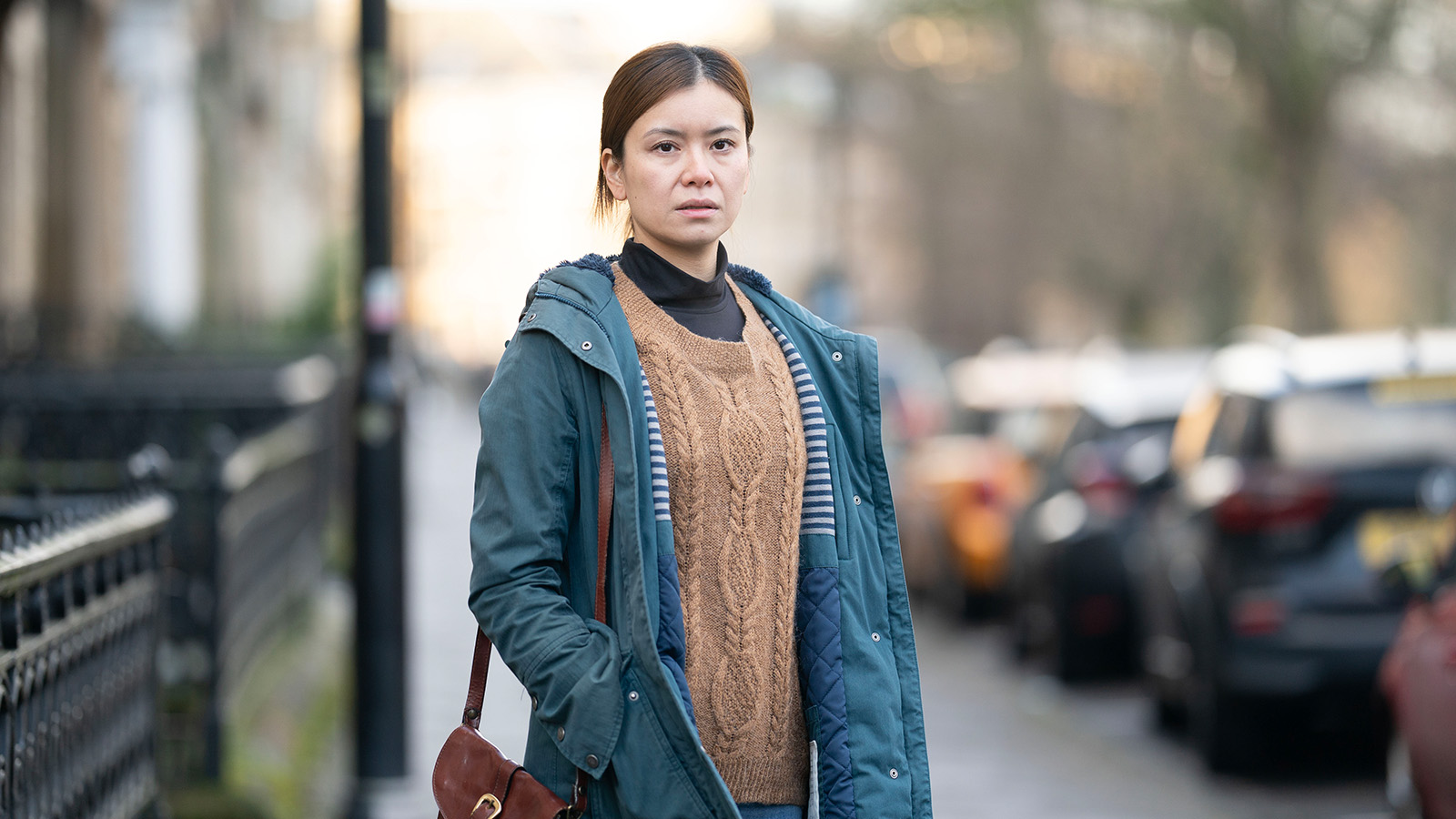 A woman (Katie Leung) stands on a city street, wearing a blue jacket over a brown sweater. She has a serious expression and a cross-body bag. Parked cars line the blurred background.