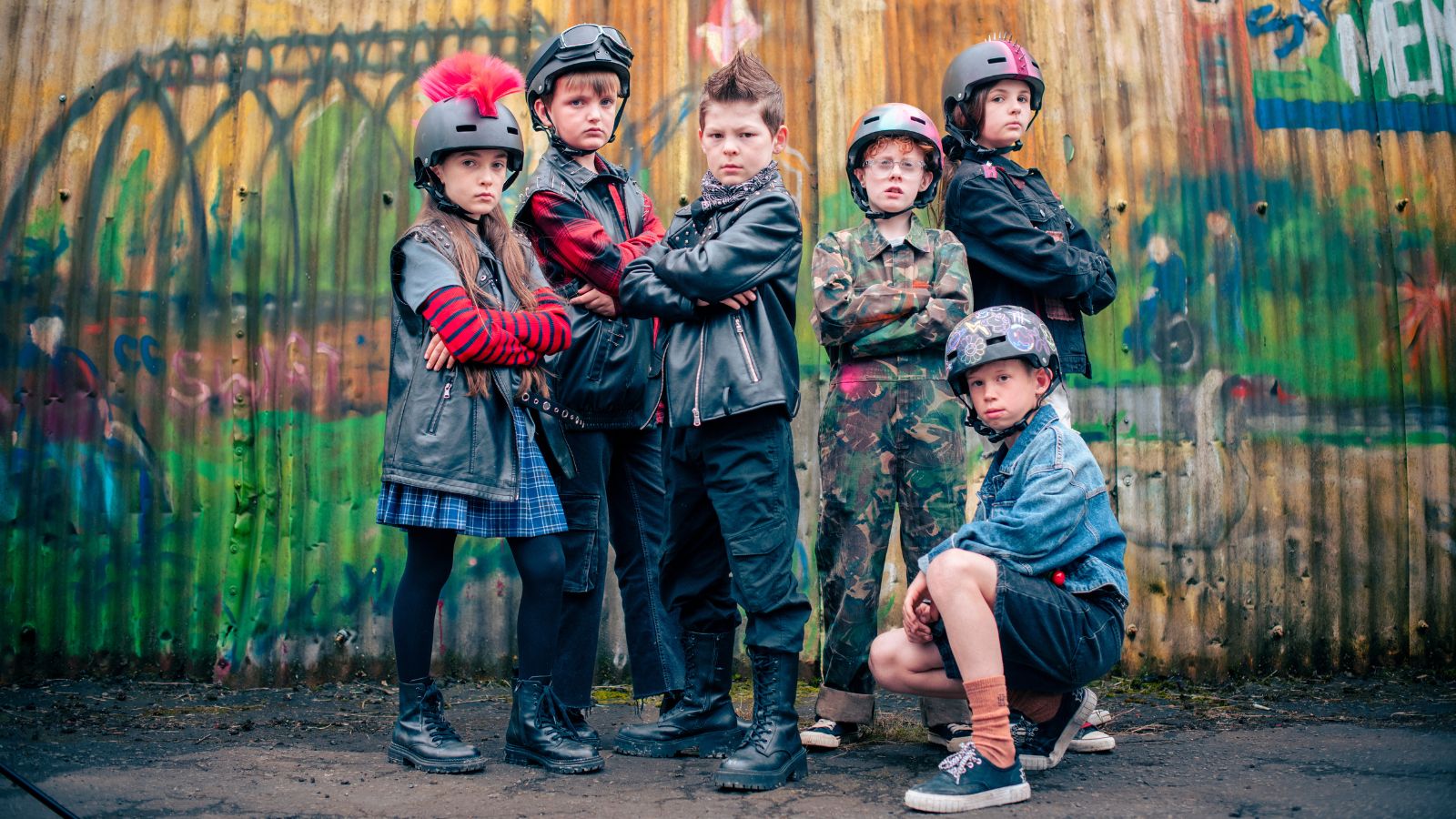 Six children stand in front of a garage door covered in graffiti. They wear a range of punkesque clothing and headwear.