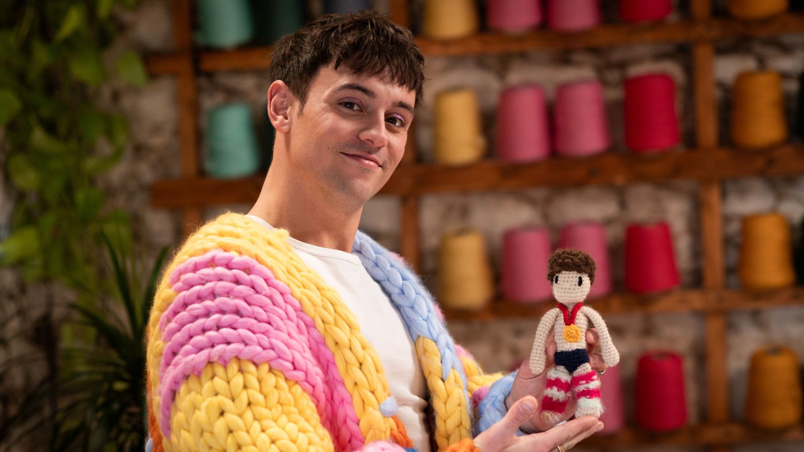 A person wearing a chunky knit sweater in pastel colours holds a small crocheted doll. The background features shelves filled with colourful yarn spools.
