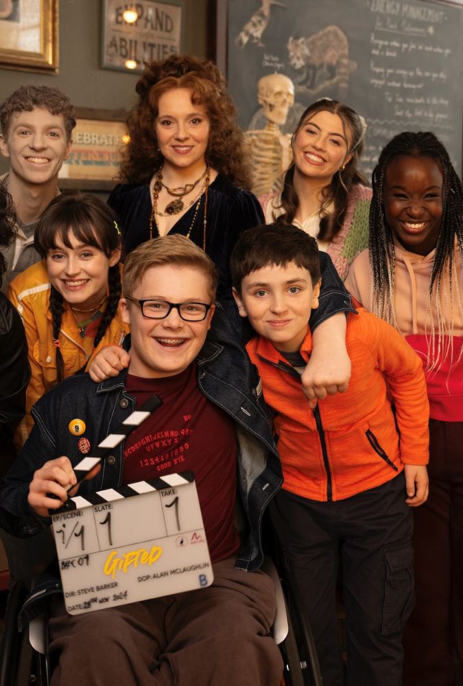 Promotional image from Gifted, which shows a group of teenagers standing together smiling at the camera. A boy in the centre holds a clapper board.