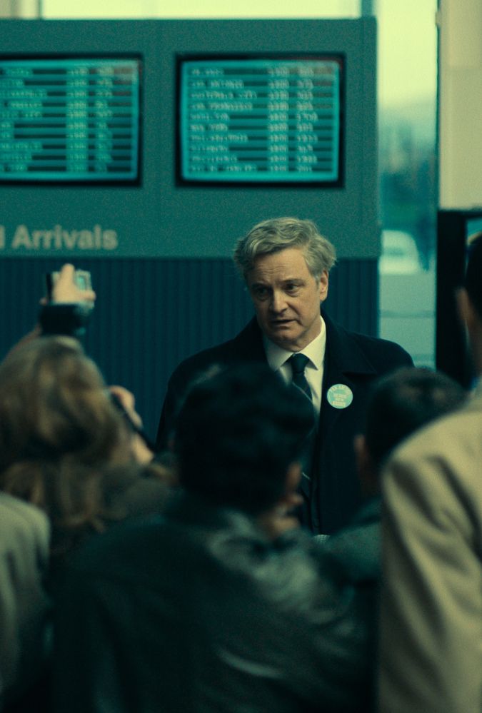 Still from Lockerbie, showing Colin Firth in a suit speaking to a crowd of journalists. He stands in front of an arrival and departures board.