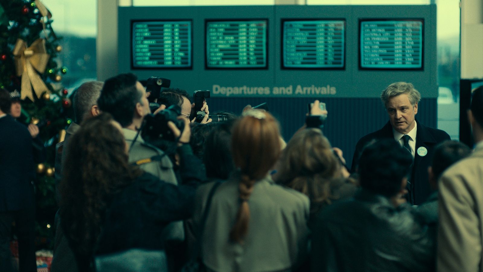 Still from Lockerbie, showing Colin Firth in a suit speaking to a crowd of journalists. He stands in front of an arrival and departures board.