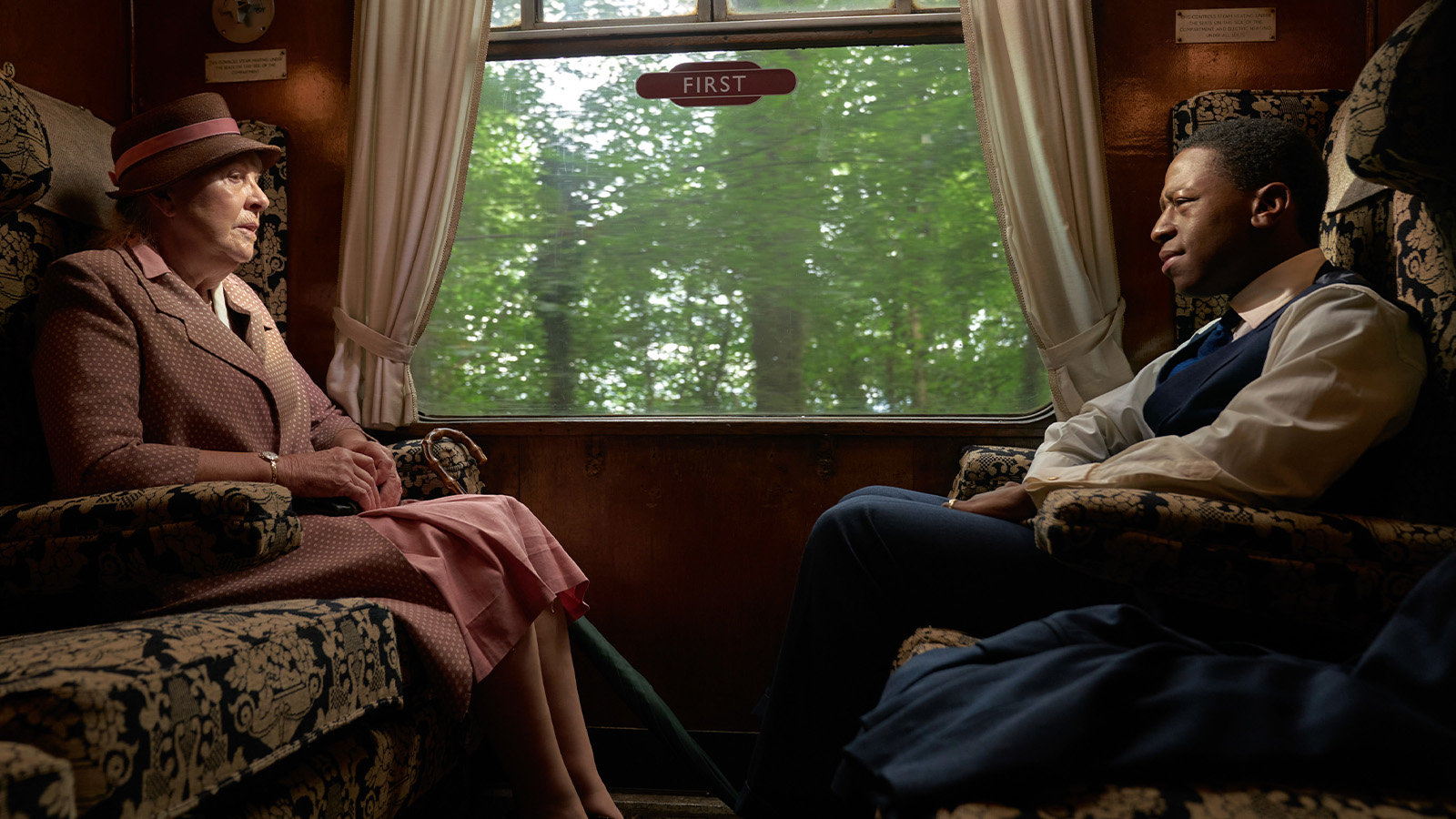 Two people sitting across from each other on a train - one is a man wearing a 1940s style black suit with hat, the other an older lady