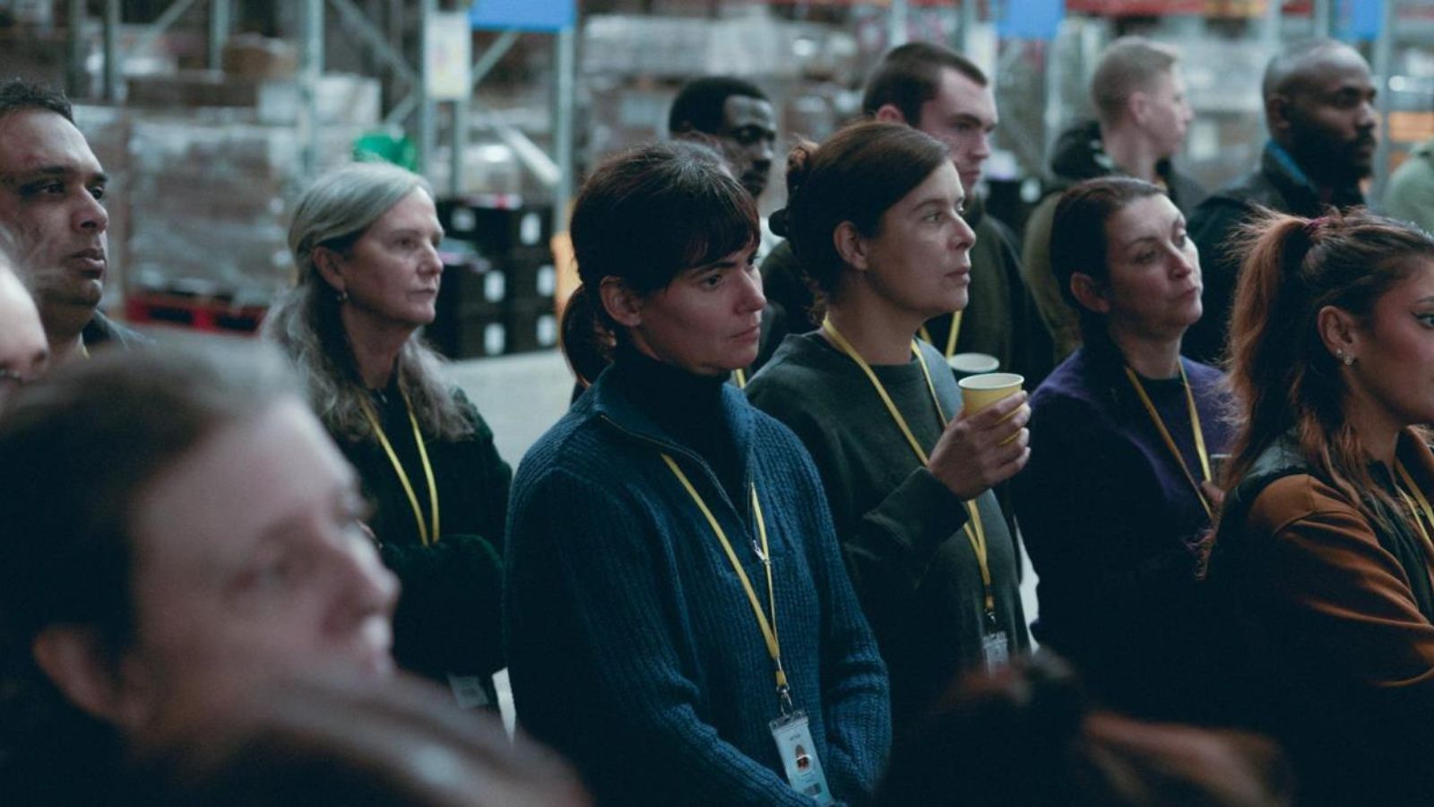 Still from On Falling which shows a groups of people standing together in a warehouse. They wear lanyards and are listening to someone speak.