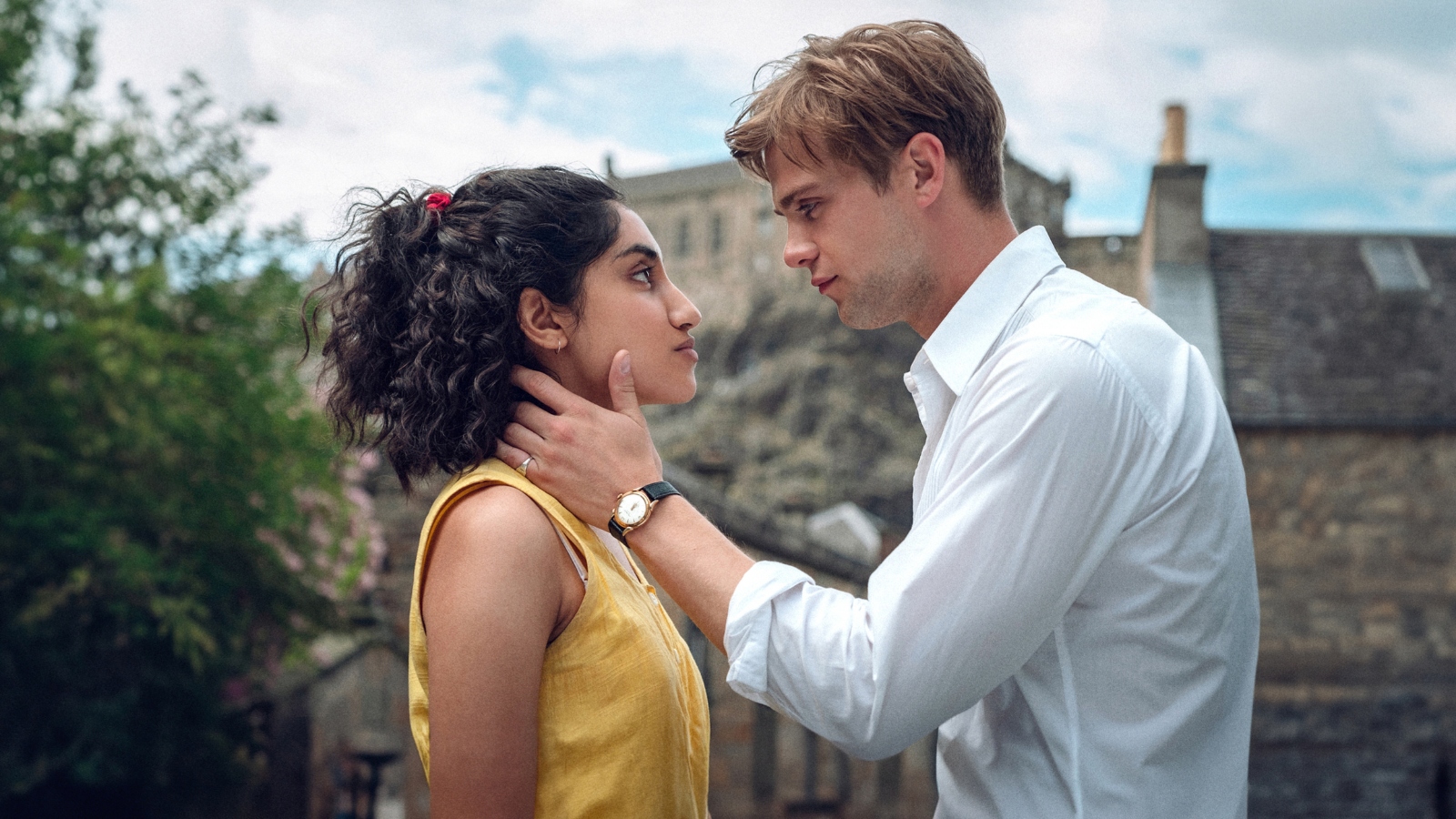 The still from Netflix production One Day, features a man, Dexter played by actor Leo Woodall and a woman, Emma, played by actress Ambika Mod, looking at each other, with Dexter's arm caressing Emma's neck and chin. Behind them is part of Edinburgh Castle.