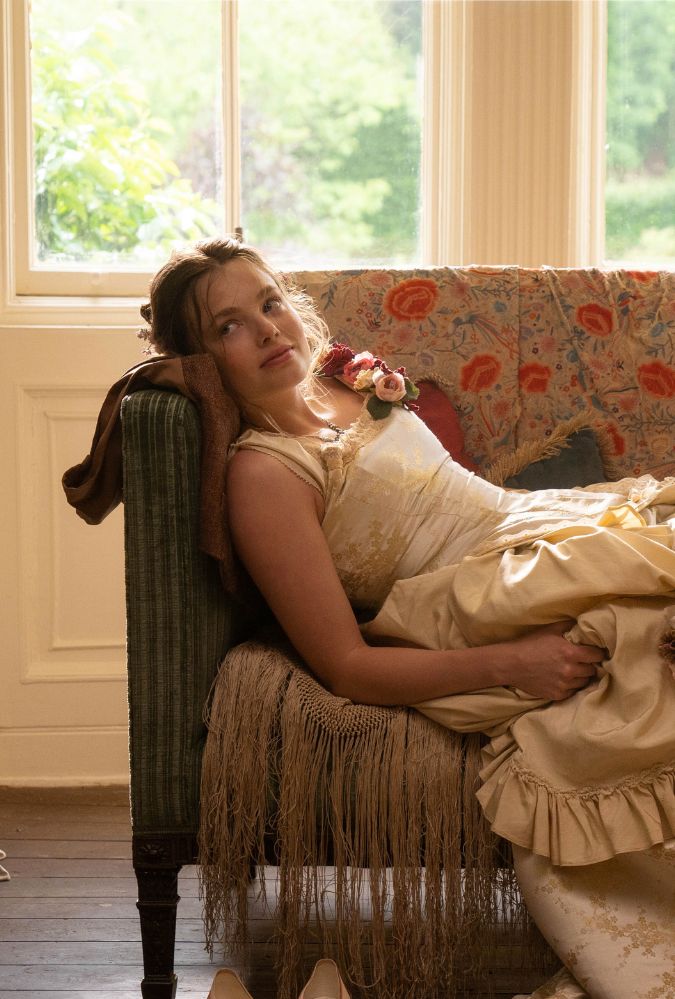 Image shows actor Kristine Frøseth lying on a sofa in a brightly lit room. She wears a cream coloured 1870s style dress.