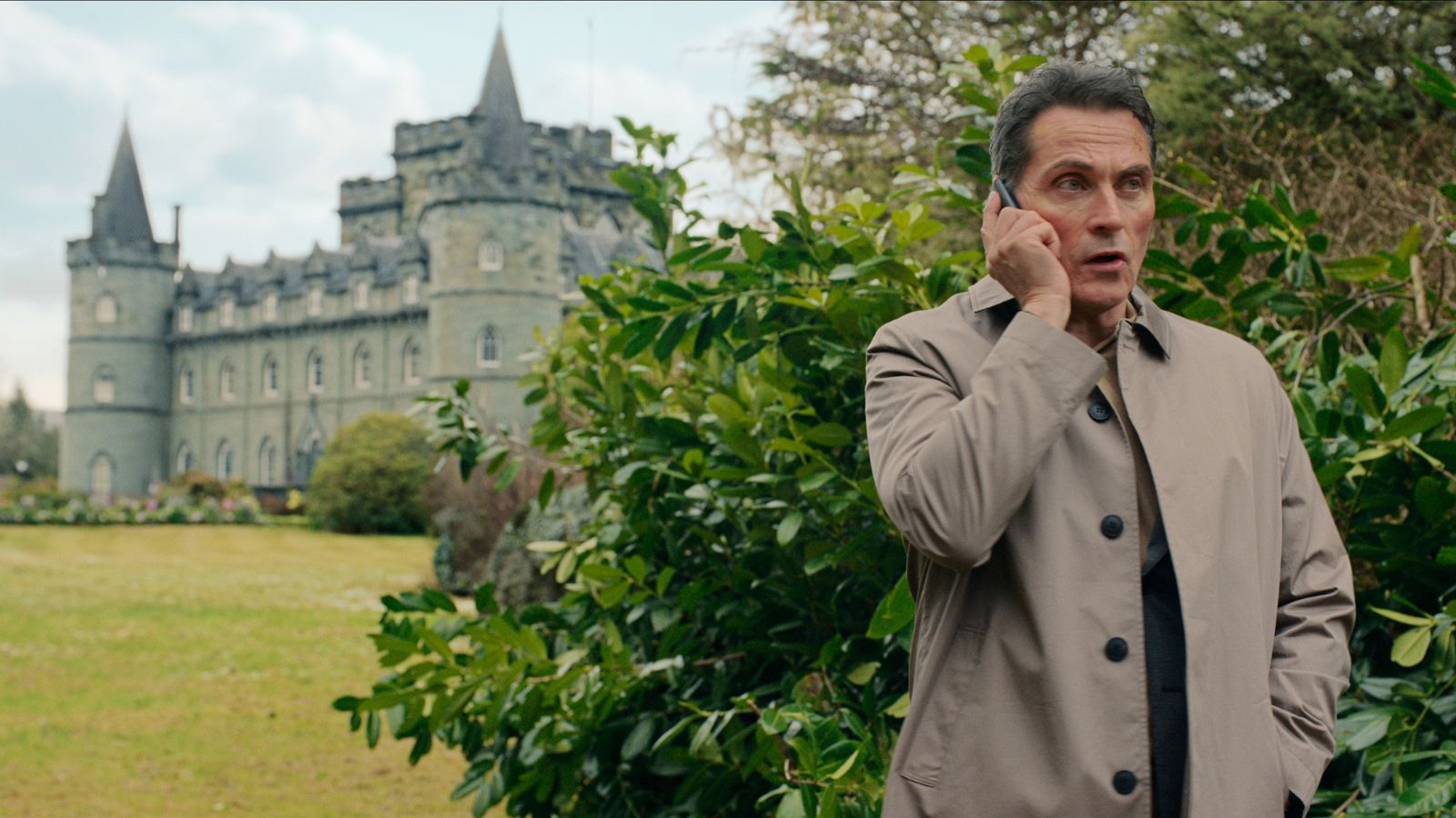 Still from The Diplomat series 2, showing Rufus Sewell standing outdoors in front of Inverary Castle on a mobile phone. He wears a long brown jacket.