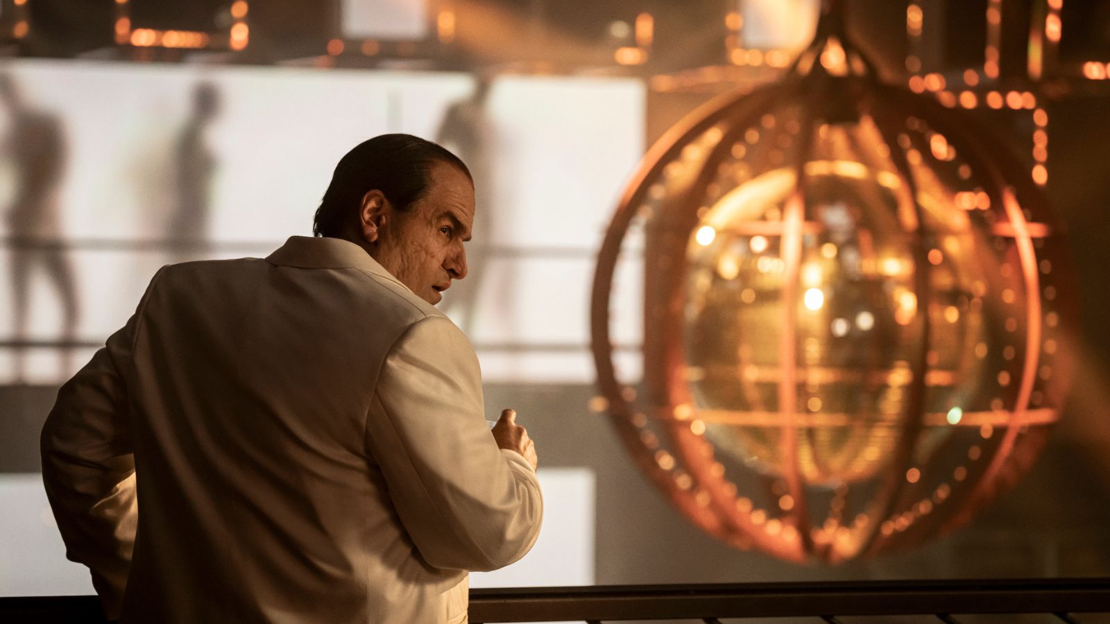 Still from The Penguin, showing a close up of actor Colin Farrell as Oswald Cobblepot. He wears a white suit jacket and has his back to the camera, turning to the right. He is standing indoors on a balcony, with a large gold glitter ball handing to his right.