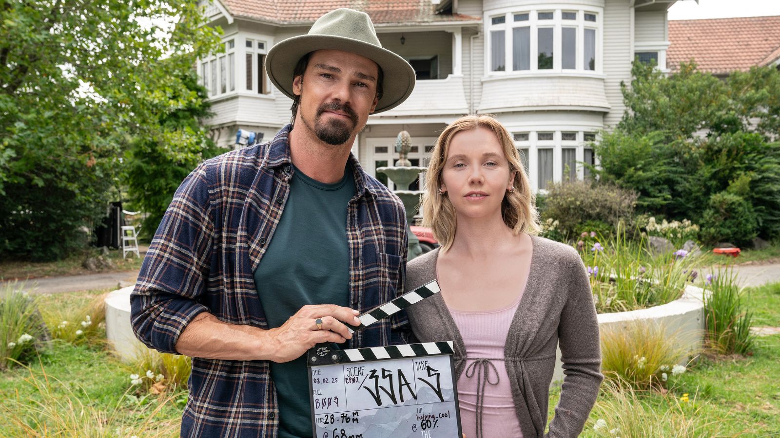 Image from The Ridge which shows actor Jay Ryan standing outside next to actor Lauren Lyle in front of a white house and garden. Jay wears a hat and holds a clapper board.