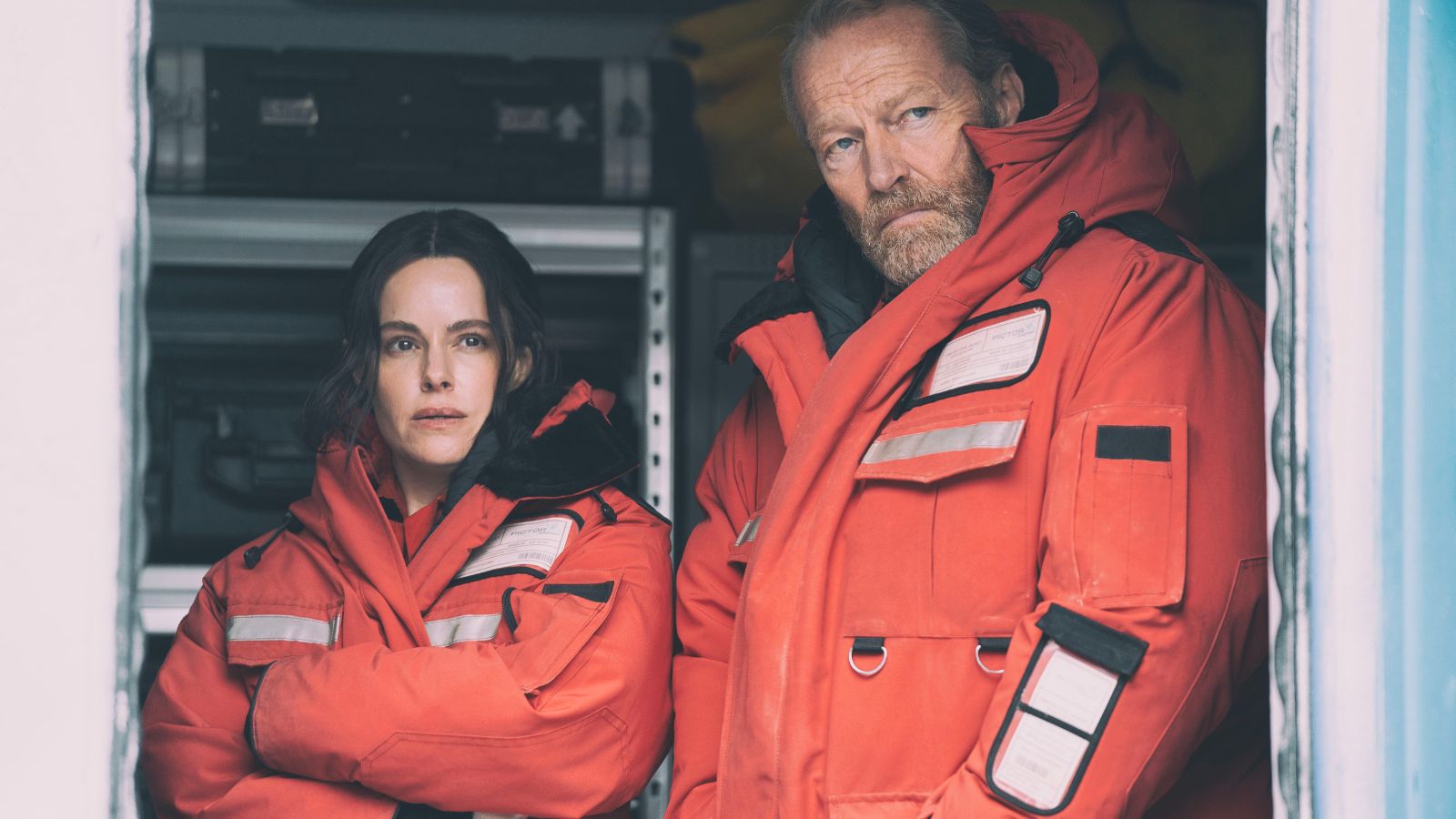 Still from the Rig season 2 showing Emily Hampshire and Iain Glen standing side by side wearing orange boiler suits.