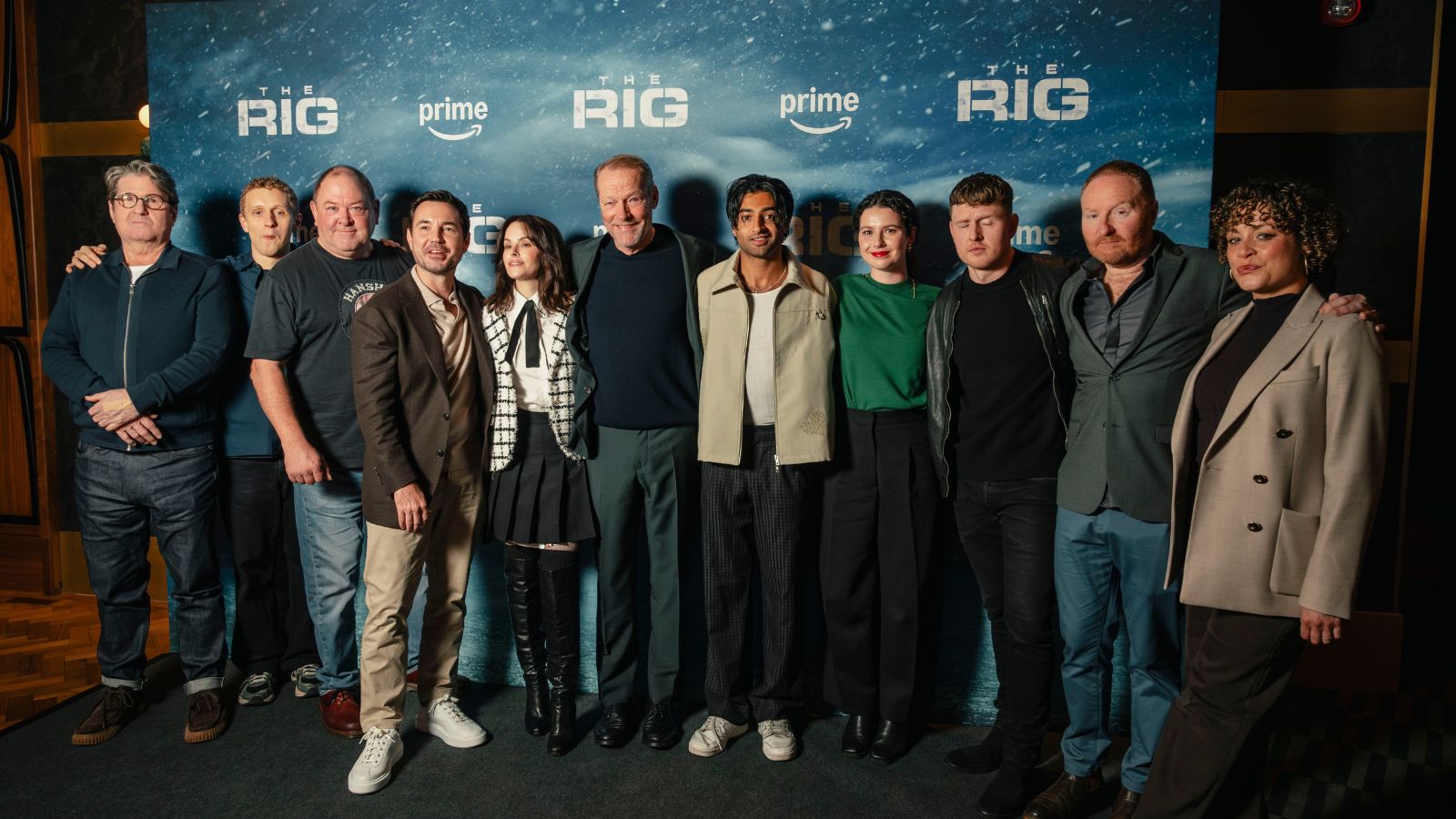 Promotional image from The Rig series 2 launch event, showing 11 cast members, including Martin Compston and Iain Glen, standing in a line in front of a board with the shows name on it.