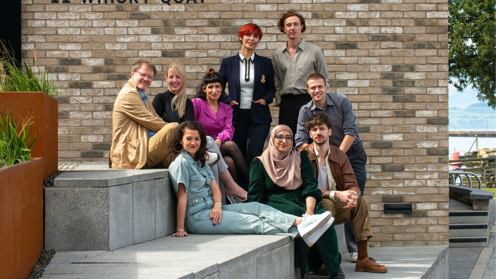 Director John Mclean (seated far left) sits with eight of the Rising Stars Scotland on the steps outside Leith Distillery in Edinburgh. They all look towards the camera and smile.