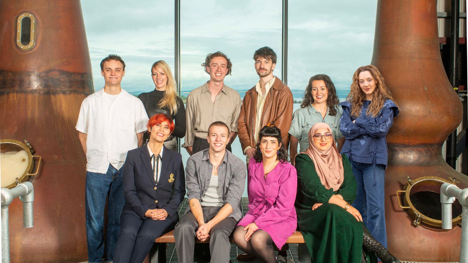 Credit: Rob McDougall/Screen International. The Rising Stars Scotland 2024 cohort are shown in a distillery in two rows. They are Standing (l-r): Séamus McLean Ross (actor), Simone Smith (writer/director), Jamie Fraser (writer/director), Sean Lìonadh (writer/director), Anna Russell-Martin (actor), Mirren Mack (actor)  Sitting (l-r): Nadira Murray (producer), Ruaridh Mollica (actor), Maryam Hamidi (writer), Raisah Ahmed (writer/director)