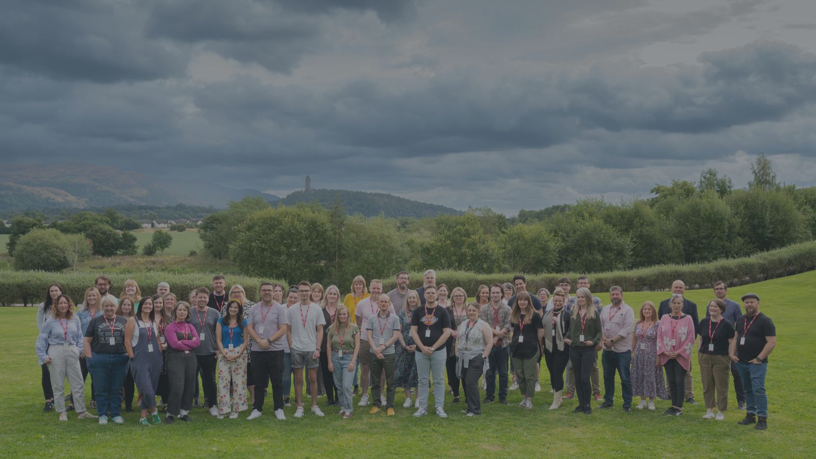 Credit Julie Howden. A group of 51 people stand in two rows outdoors on grass. In the background there are trees and in the distance the Wallace Monument.