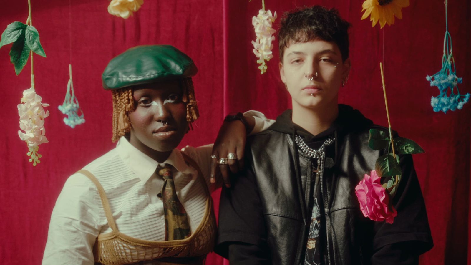 Still from Twenty Something. Two people stand in a room in front of a red background, with dried flowers hanging from above.