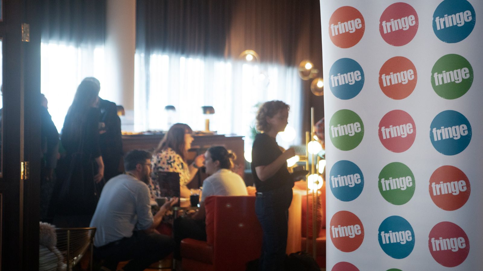 A group of people sit in together chatting in a room at a networking event. To the right is a pull up banner which has the Edinburgh Fringe logo on in in multiple colours.